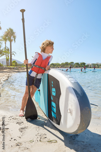 Vertical photo of a blond boy with curly hair looking to a sea with distracted expression while wearing a vest leaning against a paddle surfboard and holding the paddle stick on the beach photo