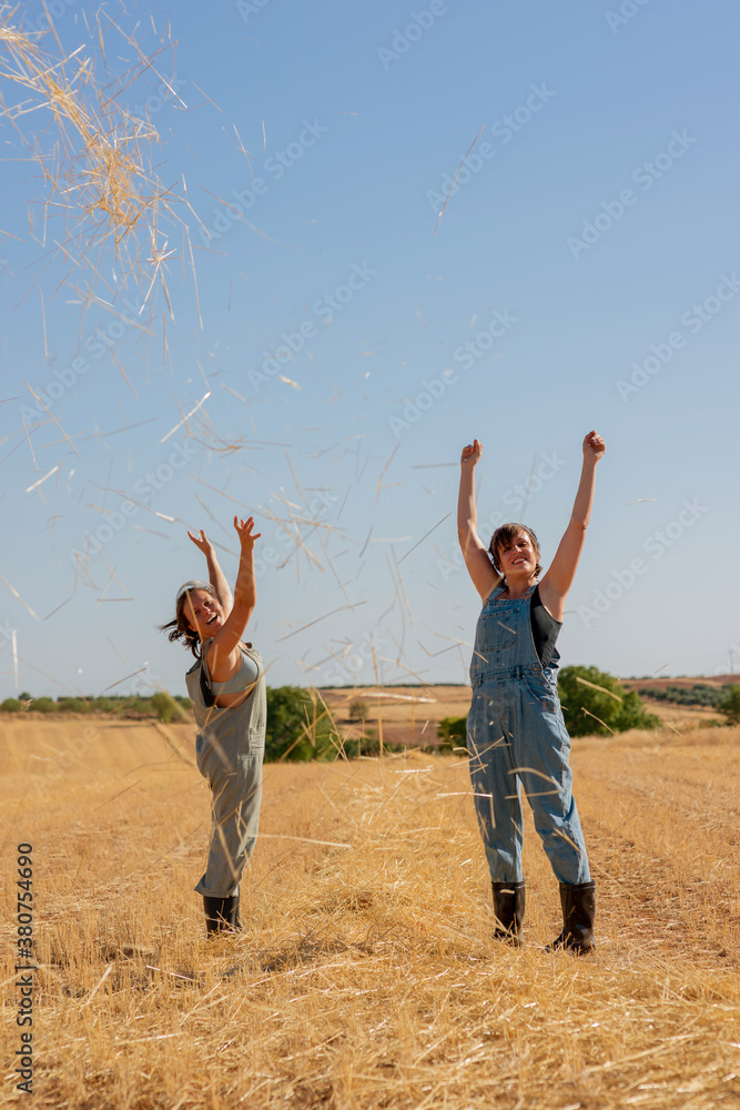 Delighted female friends standing in dry field and throwing hay in air while having fun and looking at camera Delighted female friends standing in dry field and throwing hay in air while having fun and looking at camera
