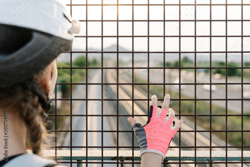 Back view of crop unrecognizable female bicyclist in protective helmet and gloves standing near grate fence on bridge over road with blurred hills in background photo