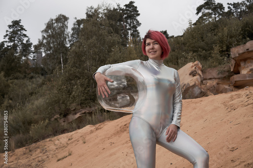 Low angle of positive young female with dyed red hair wearing silver space suit and glass helmet and looking away while walking on rocky formation photo