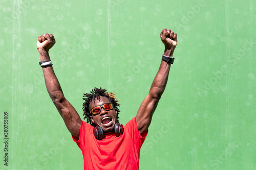 Delighted African American male in sunglasses and with headphones celebrating goal achievement with raised arms and fists up