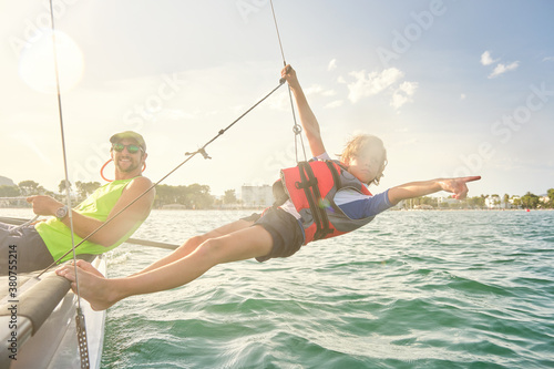 Blond boy with a life jacket attached to a rope from a boat hanging out to sea pointing to the horizon with a man next to him sitting on the boat steering the wheel with the sun illuminating them