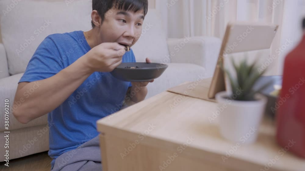 Asian young man enjoys watching a tablet while eating a spoon of riceberry in cozy living room, people and daily routine, having a meal, nutrition and delicious recipe, self entertainment at home Asian young man enjoys watching a tablet while eating a spoon of riceberry in cozy living room, people and daily routine, having a meal, nutrition and delicious recipe, self entertainment at home