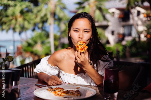 Happy asian pretty woman hungry having pizza at sunny day, sunset light in outdoor restaurant. Female enjoying  food, having fun at lunch. Tropical background.  