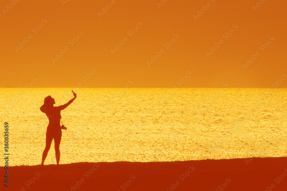 Silhouette of a woman holding a phone and taking selfie outside during sunset. Black silhouette over sunset. Woman posing on field and taking a selfie. Copy space. High quality photo