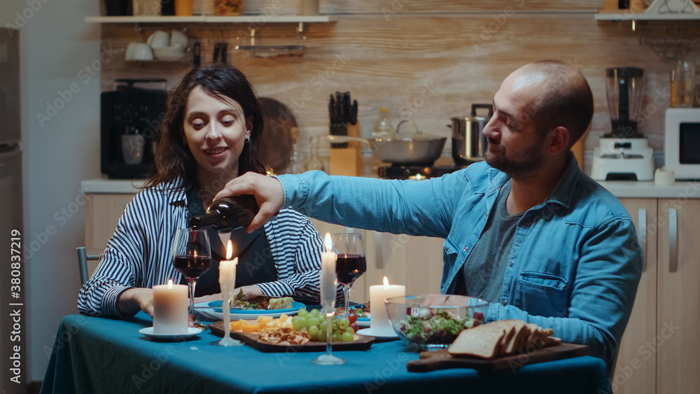 Fototapeta premium Relaxed young husband talking with his wife and pouring red wine in glasses. Romantic caucasian happy couple sitting at the table in kitchen celebrating marriage at candle lights, love and anniversary