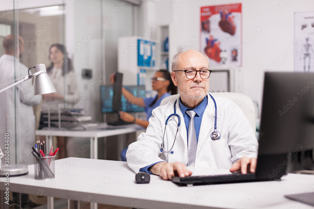 Elderly aged physician with stethoscope working on computer in clinic office while young medic is discussing illness rapport with patient on hospital corridor and nurse is holding sick man x-ray. Elderly aged physician with stethoscope working on computer in clinic office while young medic is discussing illness rapport with patient on hospital corridor and nurse is holding sick man x-ray.