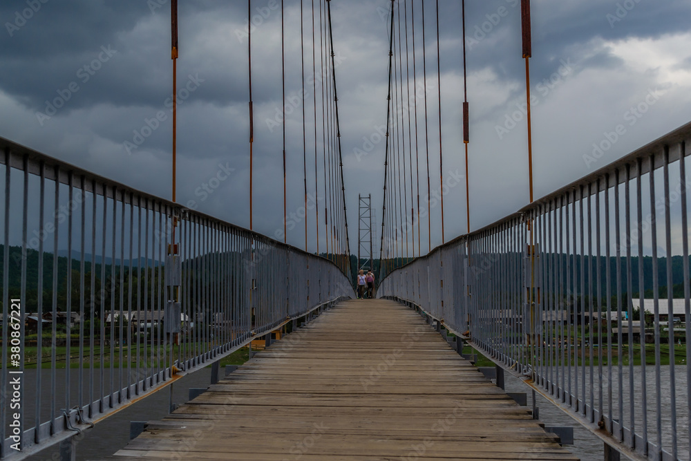 suspended pedestrian bridge with metal vertical cables and wooden path against the backdrop of village of Shamanka among Siberian mountains, cloudy landscape, geometric, symmetrical view