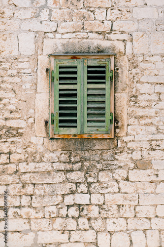 A window with closed, old, green, wooden shutters in the brick wall of the house.