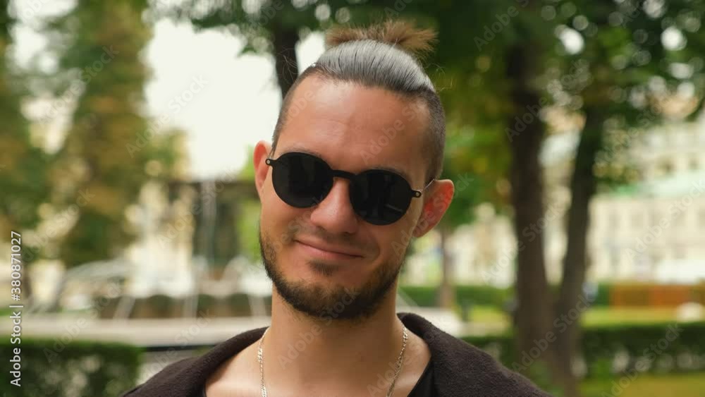 A young, Caucasian, charismatic brunette man in dark clothes and glasses with light lenses puts on dark lenses and smiles. Close-up of the face. Small ponytail on dark hair. A young, Caucasian, charismatic brunette man in dark clothes and glasses with light lenses puts on dark lenses and smiles. Close-up of the face. Small ponytail on dark hair.