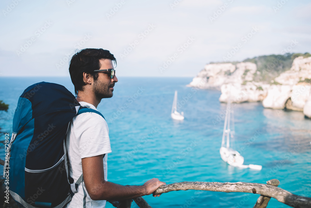 Indian male wanderer with touristic backpack enjoying scenery view from above on berth for travel yachts at Balearic islands, man exploring picturesque landscape in Menorca during adventure trip