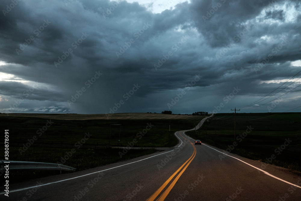 A long car drives toward a storm on a prairie road as dusk. A long car drives toward a storm on a prairie road as dusk.