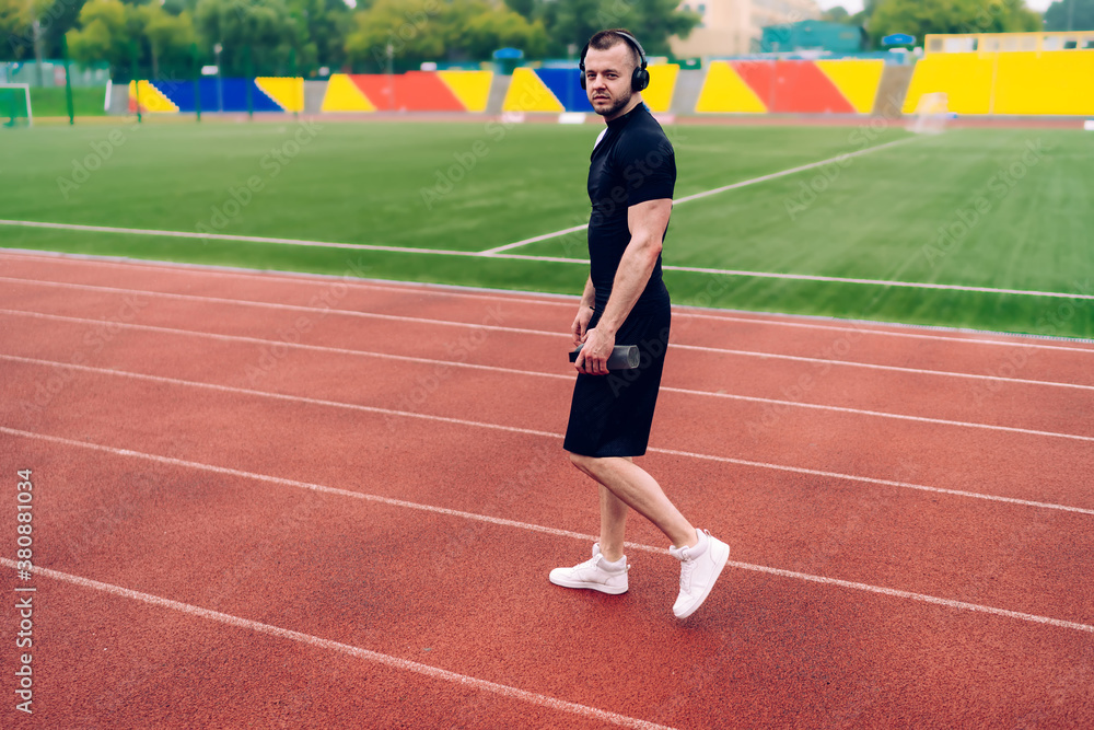 Handsome caucasian male in trendy wear and headphones walking on stadium listening favorite songs, portrait of hipster guy prepare for cardio workout enjoying online radio in sound accessory