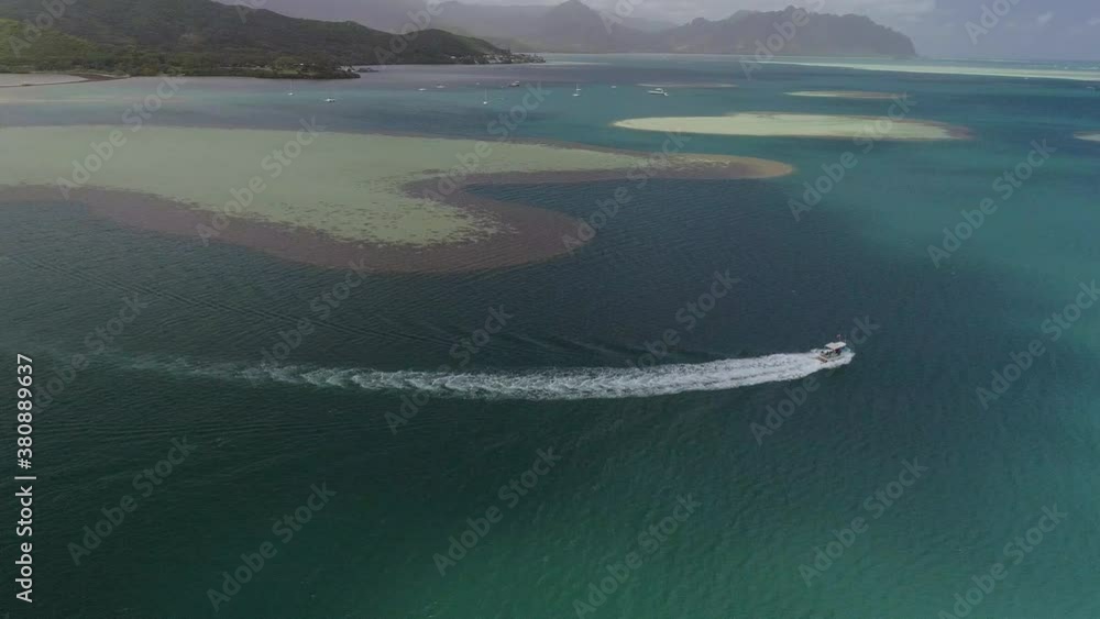 An extreme wide shot from above of a tourist boat navigating the sandy mounds of Coconut Island, Kaneohe Bay, Hawaii, and leaving a curved foamy trail in the beautiful blue tropical waters. An extreme wide shot from above of a tourist boat navigating the sandy mounds of Coconut Island, Kaneohe Bay, Hawaii, and leaving a curved foamy trail in the beautiful blue tropical waters.
