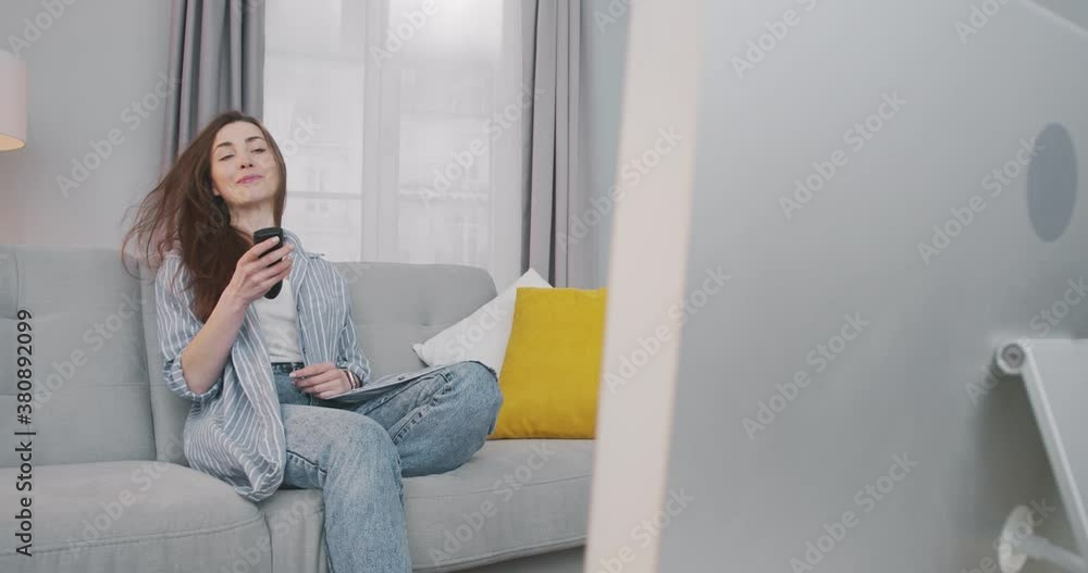 Pretty Caucasian happy young woman sitting down on sofa in living room and turn on TV. Beautiful girl in good mood changing channels with remote control while watching television at home. Rest concept Pretty Caucasian happy young woman sitting down on sofa in living room and turn on TV. Beautiful girl in good mood changing channels with remote control while watching television at home. Rest concept