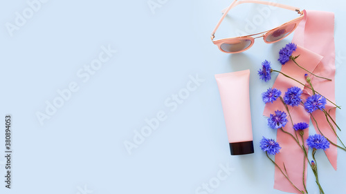 Mockup of pink squeeze bottle plastic tube with black cap, blue wild flowers, pink sunglasses and pink silk ribbon on a pastel blue background. Bottle for branding and label. Natural organic cosmetics