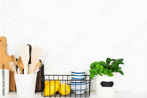 Kitchen table shelf with cutlery  spoons  spatulas  fresh basil  cutting boards  fresh vegetables  lemon on a simple wall. Horizontal  space 