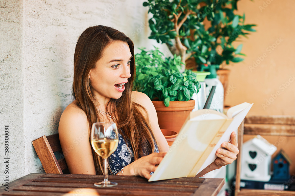 Young happy woman relaxing on the balcony, reading interesting book, drinking glass of wine Young happy woman relaxing on the balcony, reading interesting book, drinking glass of wine