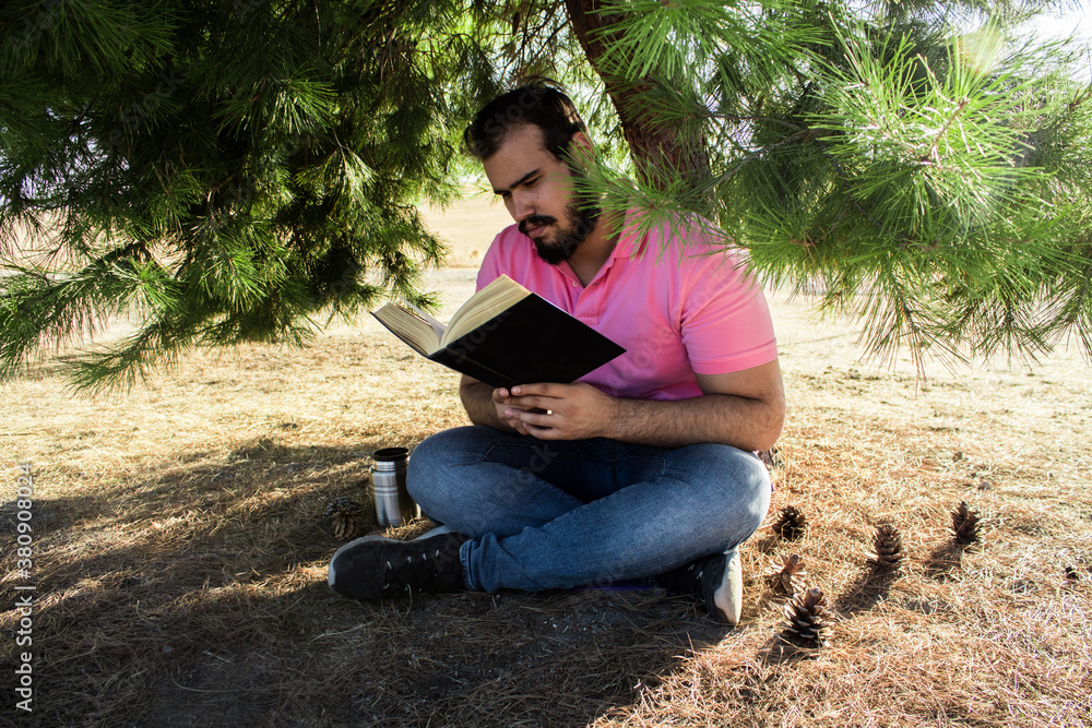 Caucasian man in jeans pants and a pink shirt sitting near a tree in a forest reading a black book Caucasian man in jeans pants and a pink shirt sitting near a tree in a forest reading a black book