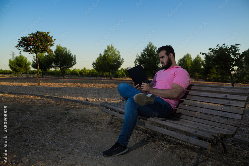 Caucasian man reading a book in the park while wearing a pink shirt and jeans