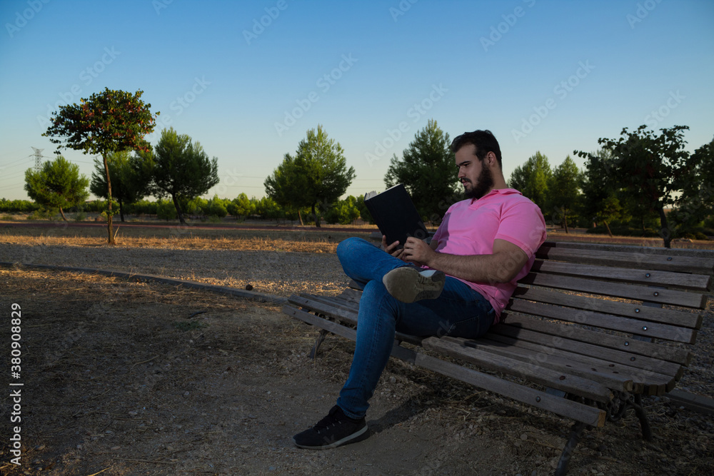 Caucasian man reading a book in the park while wearing a pink shirt and jeans Caucasian man reading a book in the park while wearing a pink shirt and jeans
