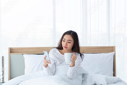 Morning girl with cup of coffee in hands in bed on a white blanket, simple, home, while playing with smartphone. Beautiful Asia woman drinking a coffee in her bed while reading from smartphone.  © Avirut S.