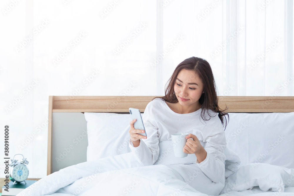 Morning girl with cup of coffee in hands in bed on a white blanket, simple, home, while playing with smartphone. Beautiful Asia woman drinking a coffee in her bed while reading from smartphone. Morning girl with cup of coffee in hands in bed on a white blanket, simple, home, while playing with smartphone. Beautiful Asia woman drinking a coffee in her bed while reading from smartphone.
