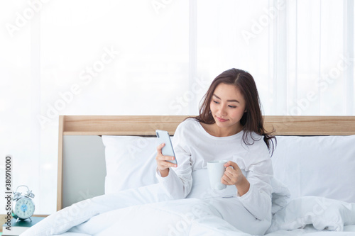 Morning girl with cup of coffee in hands in bed on a white blanket, simple, home, while playing with smartphone. Beautiful Asia woman drinking a coffee in her bed while reading from smartphone. 