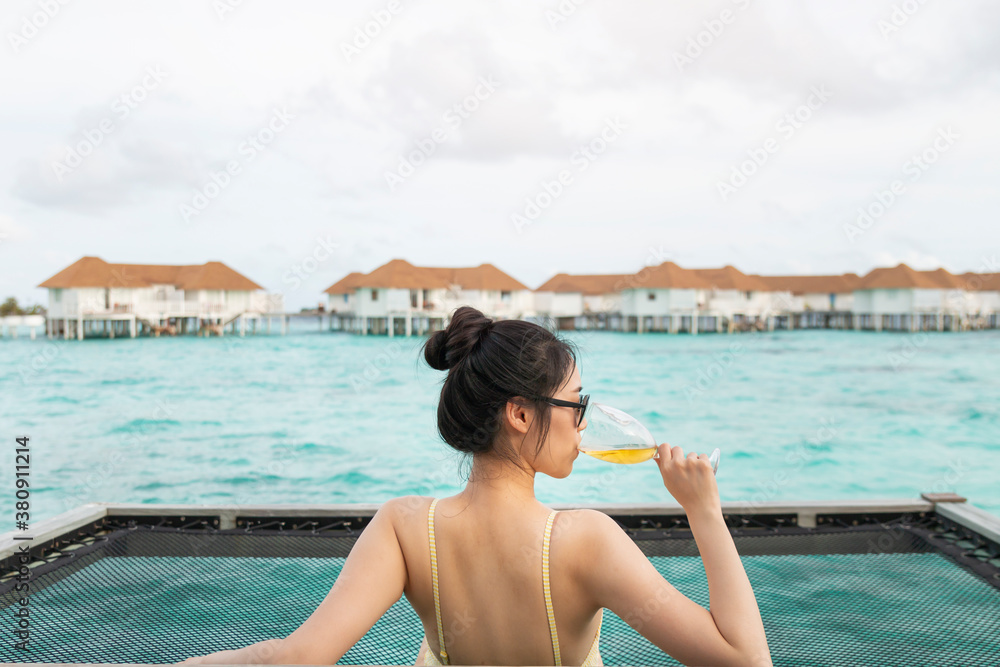 Young beautiful girl in a hammock with wine glass over the blue sea. Young girl enjoying glass of wine by the sea. Young woman watching sunset from sea hammock enjoying tropical vacations.
