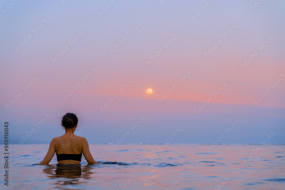 A beautiful girl bathes in the warm sea at sunset. The calm sea reflects the sunset colors of the sky. Tourist destination Bali, Indonesia. 