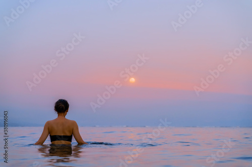 A beautiful girl bathes in the warm sea at sunset. The calm sea reflects the sunset colors of the sky. Tourist destination Bali, Indonesia.  © Konstantin