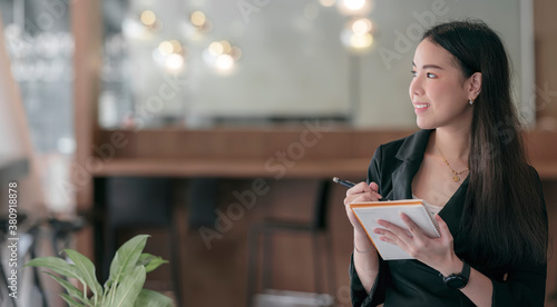 Portrait of young attractive businesswoman holding pencil and notebook while standing in modern office, copy space, panoramic view.