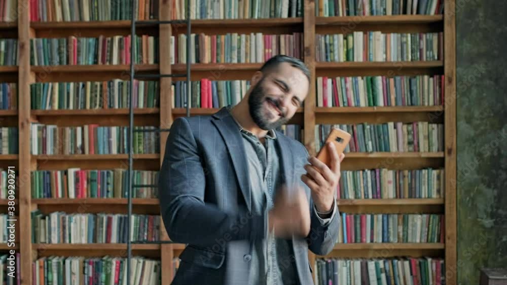 Young Man In The Library With Mobile Phone In His Hands. He Is Emotionally Playing Game On The Phone. Young Man In The Library With Mobile Phone In His Hands. He Is Emotionally Playing Game On The Phone.