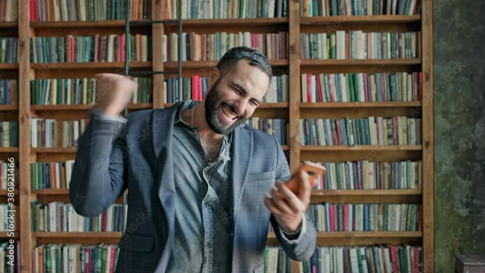 Young Man In The Library With Mobile Phone In His Hands. Something Pleasantly Surprises Him. Young Man In The Library With Mobile Phone In His Hands. Something Pleasantly Surprises Him.