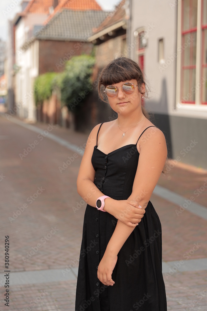 Fototapeta premium A young beautiful girl wearing in round glasses and black dress standing in city background in Holland, outdoor shoot