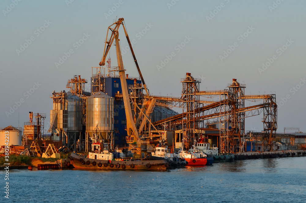 Coastal industrial factory with cranes and structures with port and transport ship. Generic view of industrial harbor with vessels docked in sunset light. Sea