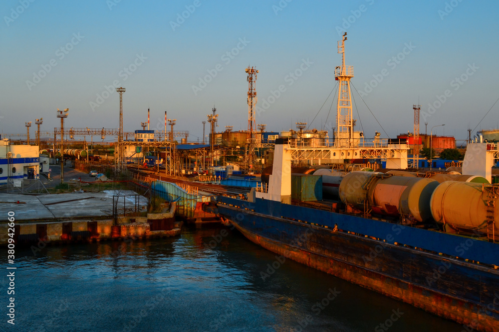 General view of an industrial harbor with moored large ships with masts, barges with cargo, boats in the red light of sunset. Blue sea, sky gradient
