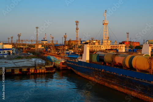 General view of an industrial harbor with moored large ships with masts, barges with cargo, boats in the red light of sunset. Blue sea, sky gradient