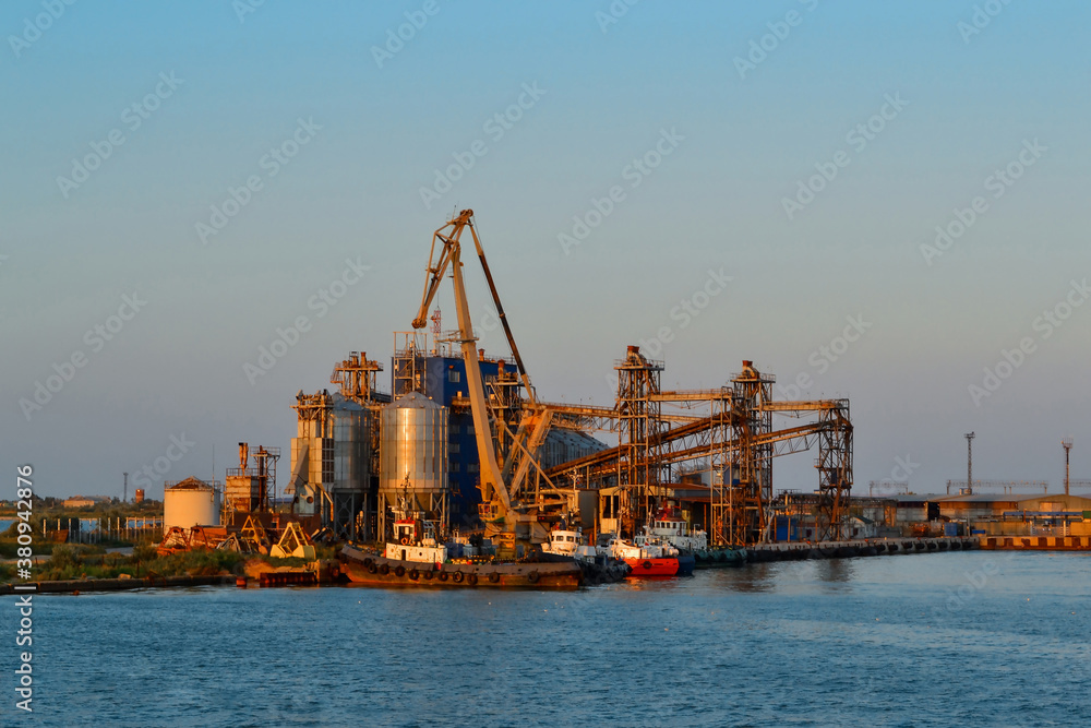 Coastal industrial seascape with red cranes and metal structures with port and transport ship. Generic view of industrial harbor with vessels docked in sunset light