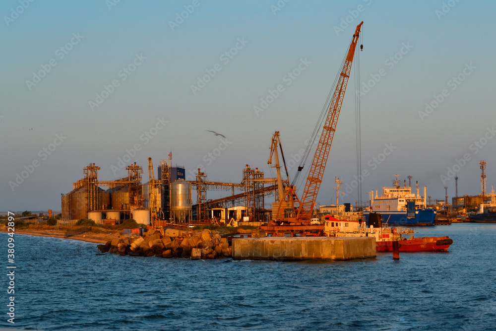 Coastal industrial factory with cranes and structures with port and transport ship. Generic view of industrial harbor with vessels docked in sunset light. Blue sea, gull