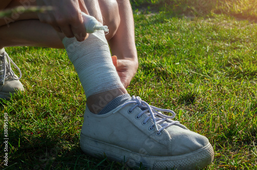 Young woman bandaging injured ankle. Injury leg while running outdoors. First aid for sprained ligament or tendon.