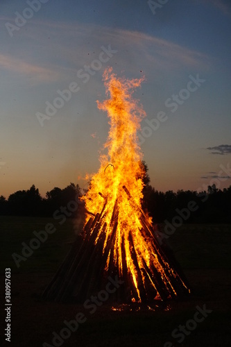 A large bonfire burns at midsummer night, with the sun setting in the background
