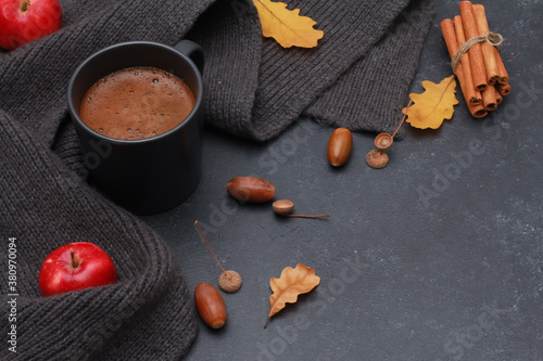 Black cup of coffee, oak leaves, acorns, red apple, grey scarf on dark background. Copy space, high angle. Autumn background, fall season, thanksgiving, coffee shop menu concept