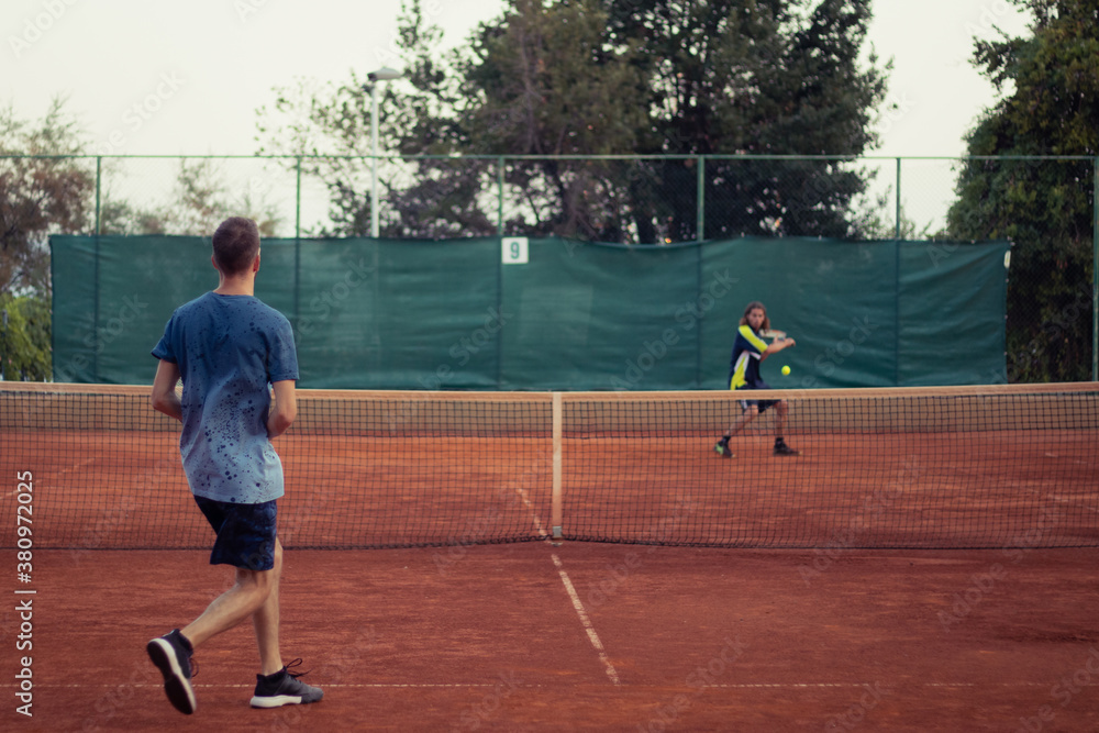 Man in blue shirt and blue pants seen from behind playing tennis, wide open clay orange playing field. Anticipating the serve from the other side, outline of the opponent hitting a ball