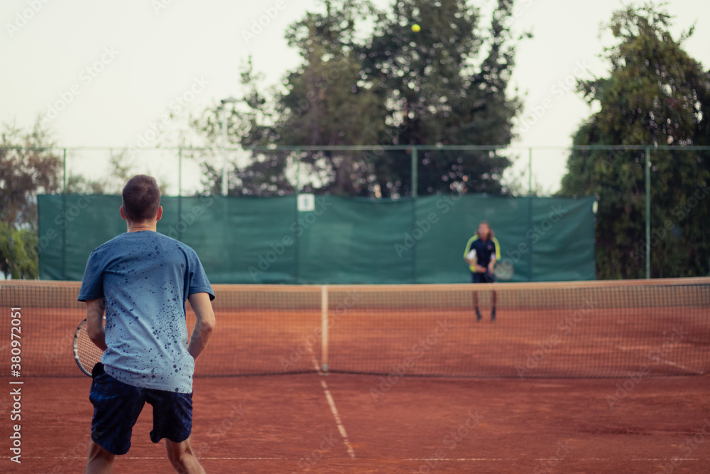 Man in blue shirt and blue pants seen from behind playing tennis, wide open clay orange playing field. Anticipating the serve from the other side, outline of the opponent hitting a ball