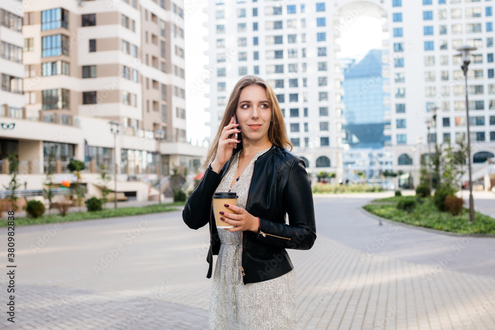 Fototapeta premium Beautiful woman is speaking on the phone with a cup of coffee at the city street background.