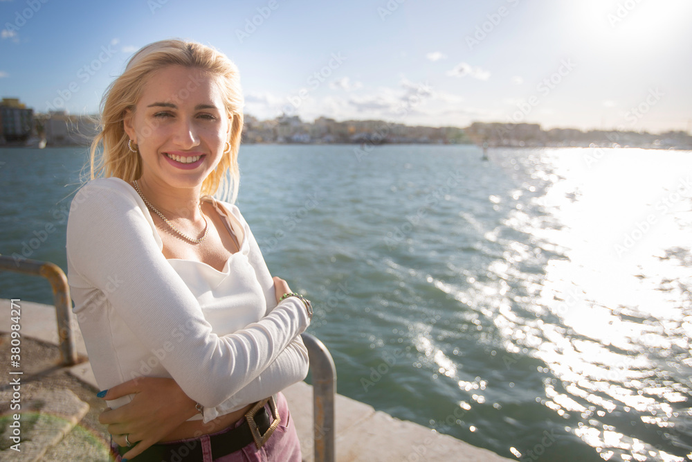 Outdoor, day. Portrait of an Italian woman with long blonde hair in a white low-cut top enriched by a delightful necklace in mother of pearl in the port