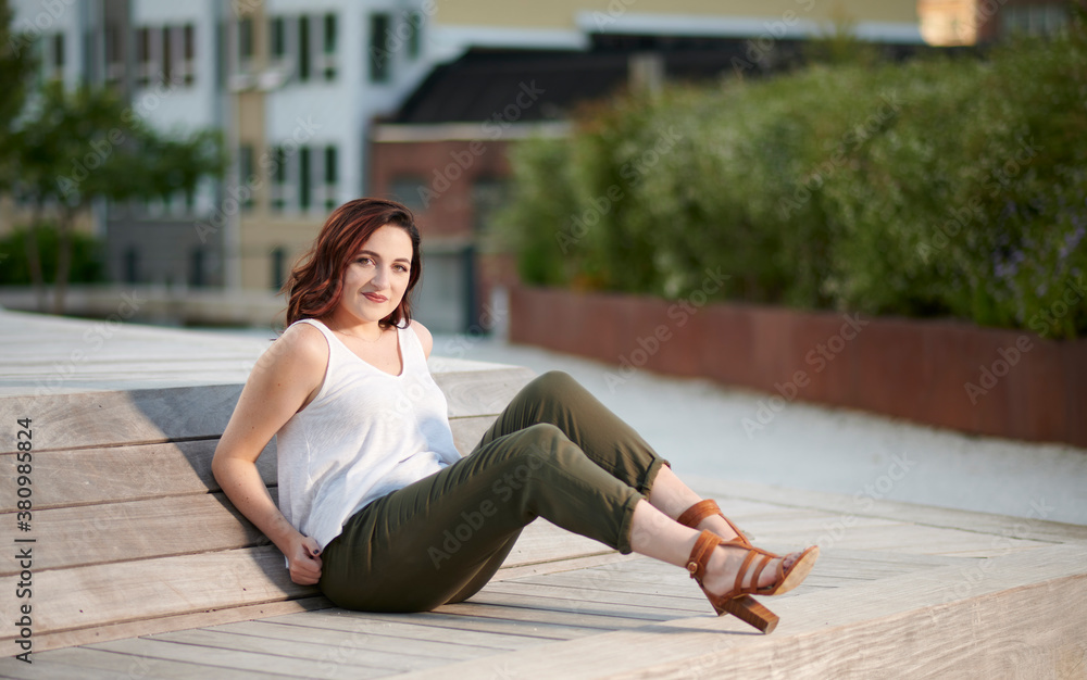 Stunning young woman poses for photo in late summer in elevated urban park - white blouse and green pants - sitting
