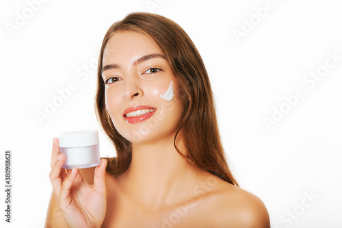 Close up image of young beautiful woman applying facial cream to face, white background, holding jar with moisturizer