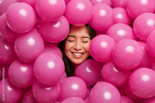 Positive cheerful young woman giggles positively with closed eyes and smiles broadly surrounded by rosy air balloons celebrates birthday spends free time on graduation party has childish mood. © wayhome.studio 